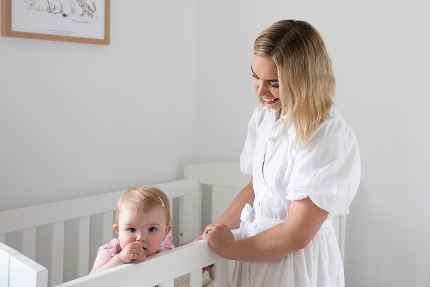 A baby standing in a cot, while her mum looks on smiling.