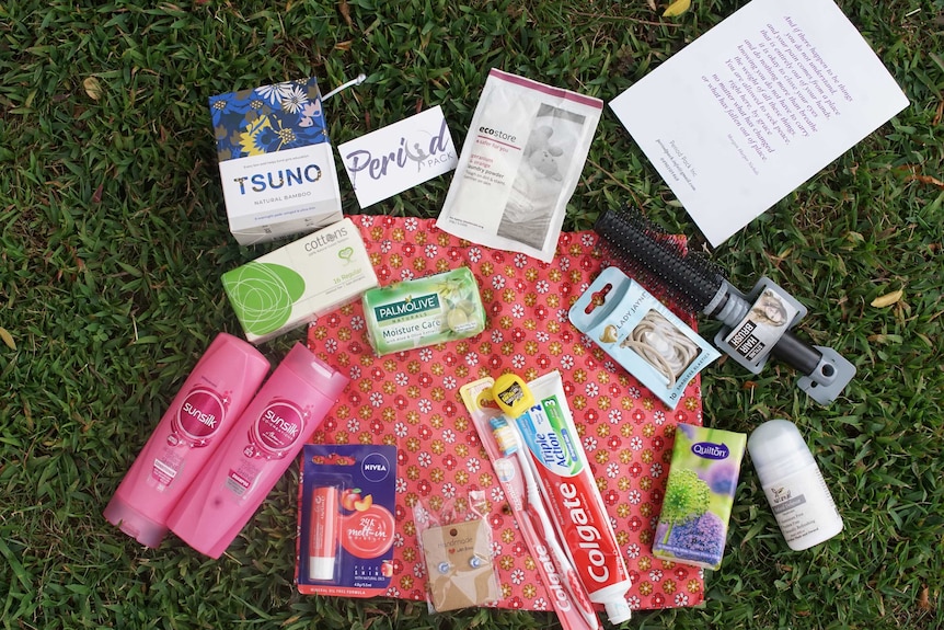 Period products and basic hygiene items are seen on top of a cloth bag resting on grass.