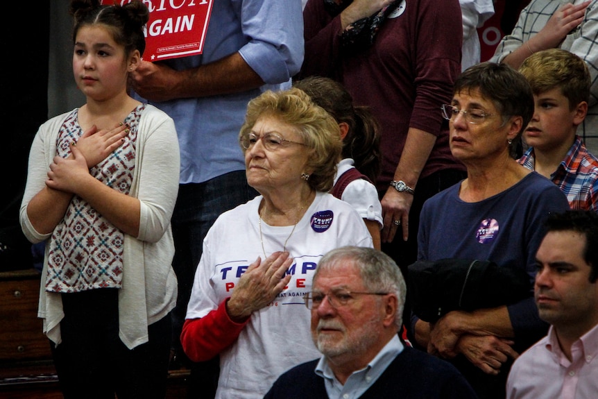 Donald Trump supporters standing up in a stadium, holding hands across their chest.