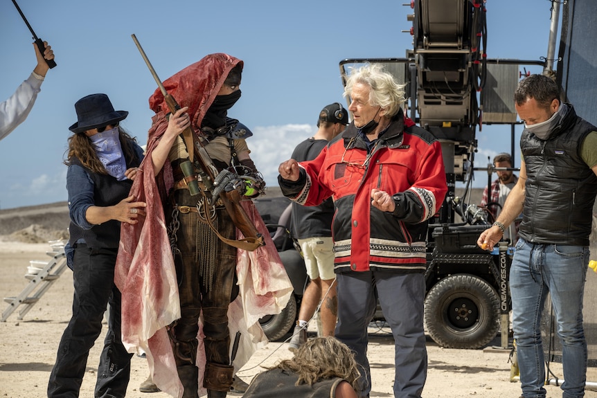 A set of masked crew assist Taylor-Joy's with her robe costume as she looks to director George Miller describing something.