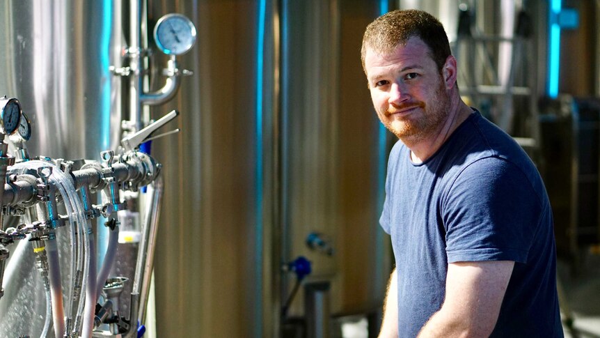 A man in a blue t-shirt looks at the camera as he adjusts hoses in a brewery.