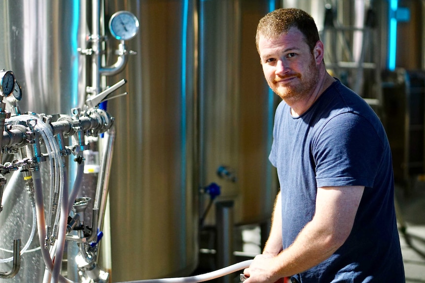 A man in a blue t-shirt looks at the camera as he adjusts hoses in a brewery.