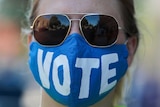 A woman wears sun glasses and a face mask that says "vote".