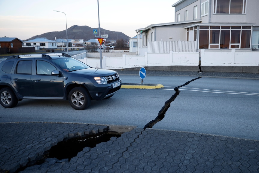 A large crack in the road is pictured with a car in front of it.