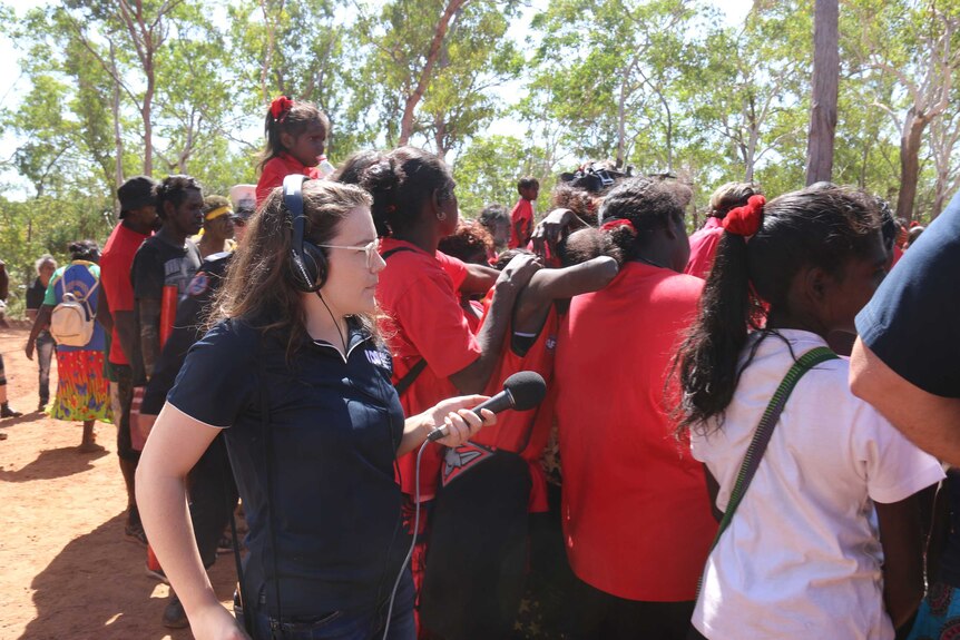 Woman with headphones on and a microphone recording a group of Indigenous people with bush in the background.