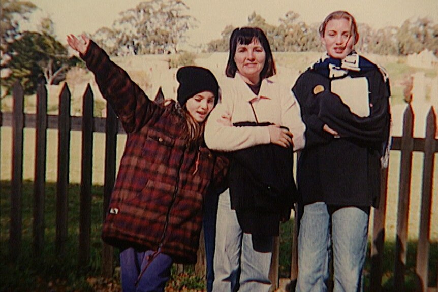 An old photo from 1996 of a young girl on the left standing next to her mother and older sister.
