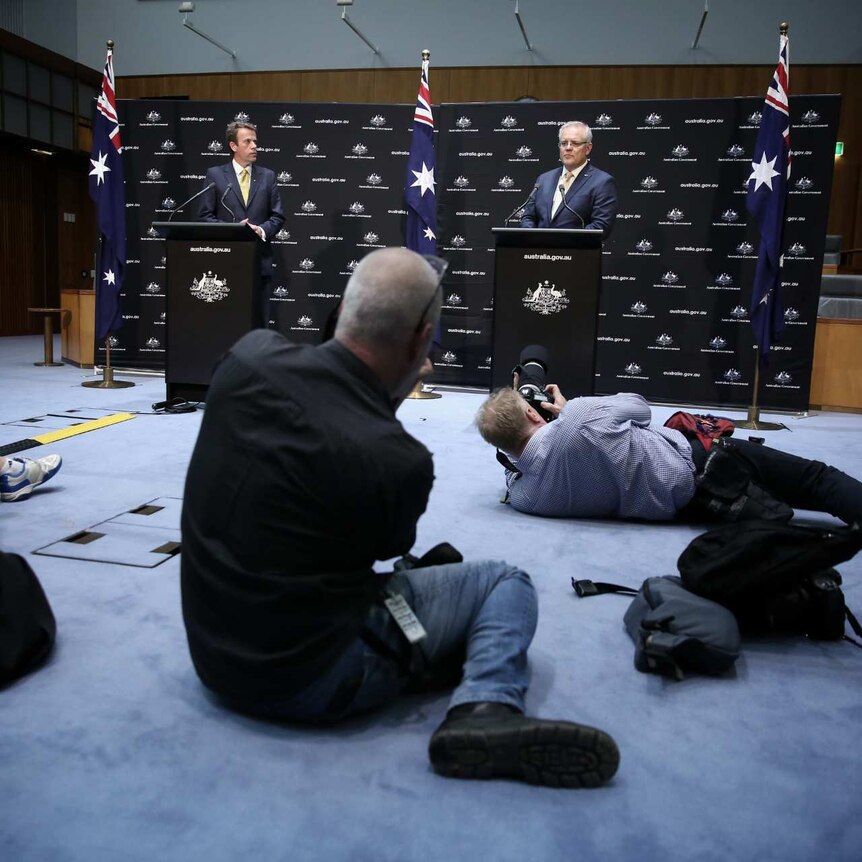 Photographers take photos of Scott Morrison and Dan Tehan, who are standing at separate podiums.