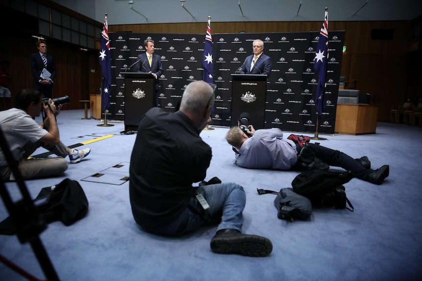Photographers take photos of Scott Morrison and Dan Tehan, who are standing at separate podiums.