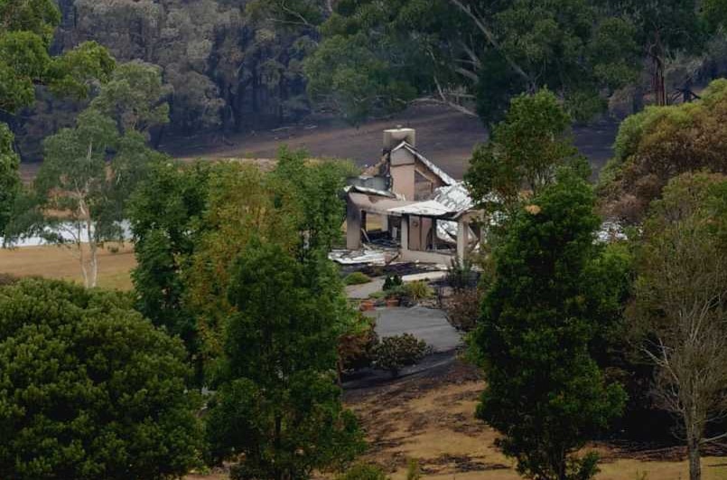 Rural wooded area with long driveaway surrounded by trees with glimpse of shell of burnt-out home