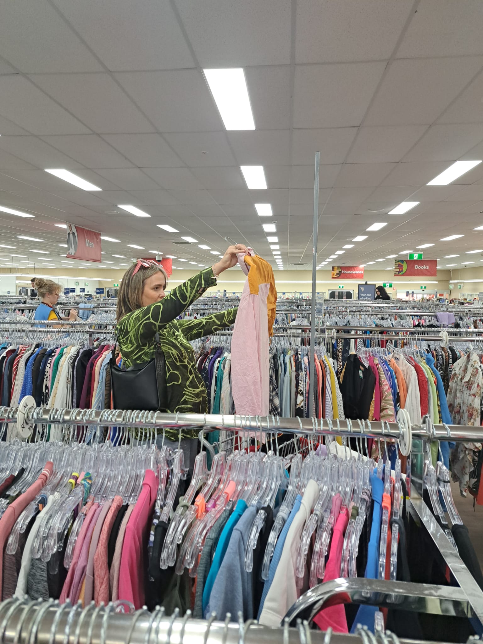 Op shopper Bec Brewin holds up a jumper to inspect while browsing a large op shop full of clothes.