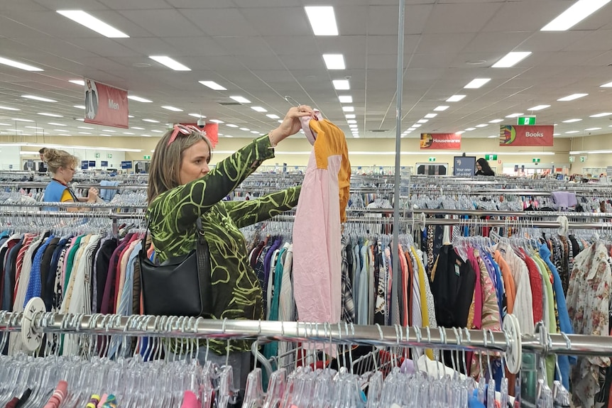 Op shopper Bec Brewin holds up a jumper to inspect while browsing a large op shop full of clothes.