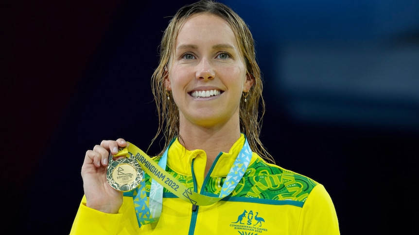 Smiling Emma McKeon in yellow tracksuit top holds gold medal next to her face