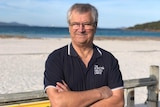 A man with short grey hair and glasses wearing a navy polo shirt standing with arms folded at the beach