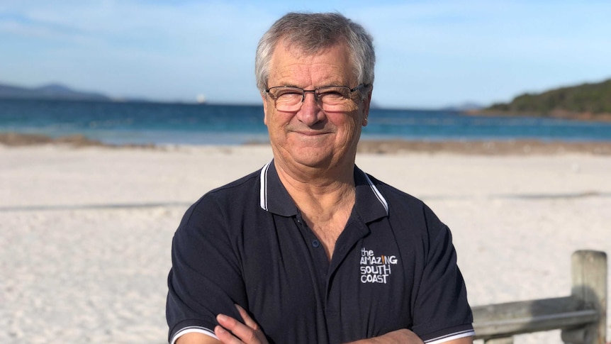 A man with short grey hair and glasses wearing a navy polo shirt standing with arms folded at the beach