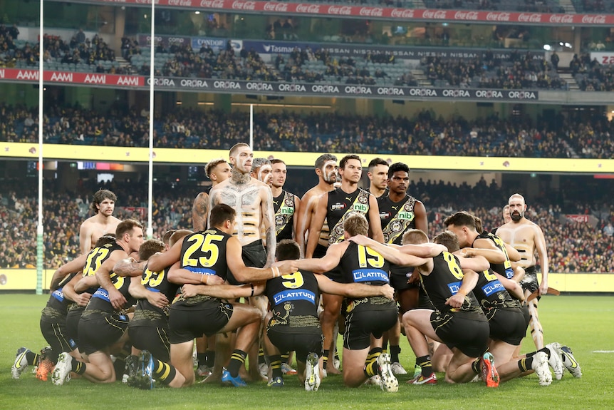 Richmond players kneel in a circle around men who stand and look out, some of whom are shirtless and have paint on them