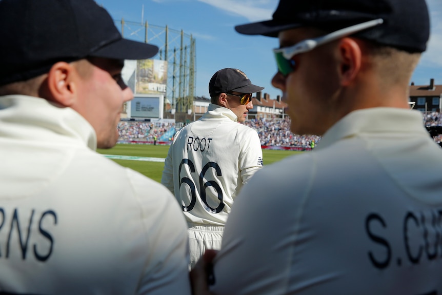 Joe Root stands on the oval cricket ground with two players framing him in the foreground