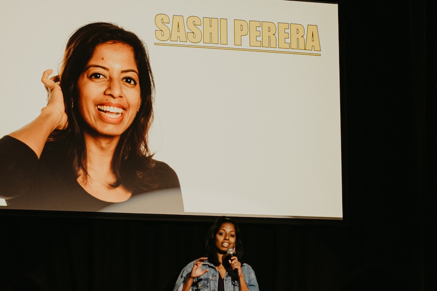 A woman standing on stage holding a microphone with her photo on screen behind her