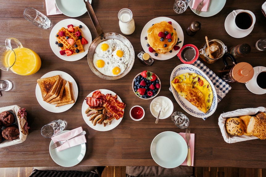 Birds-eye view of table with toast, eggs, bacon, fruits, pancakes, waffles on top.