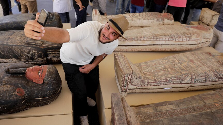 A man takes a selfie next to displayed sarcophaguses that are around 2500 years old,.