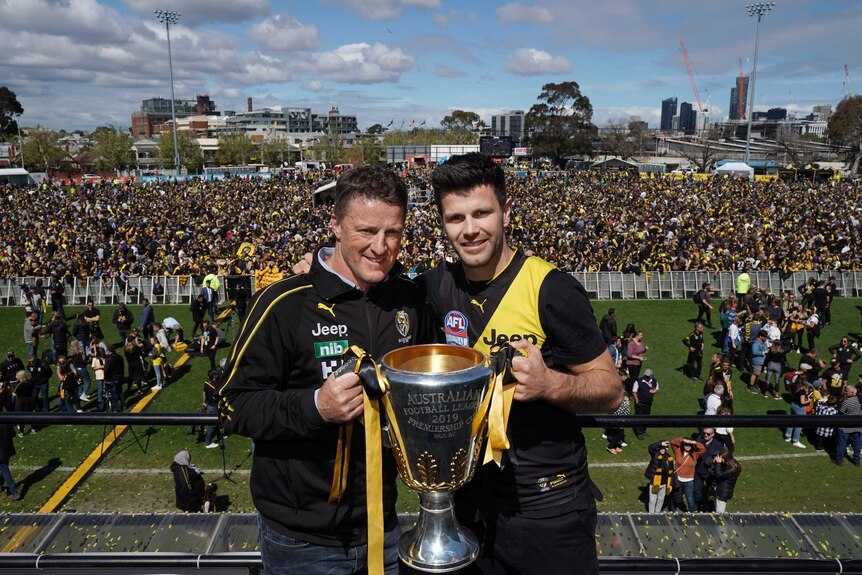 Damien Hardwick and Trent Cotchin smile happily holding the 2019 AFL premiership cup in front of a large crowd.