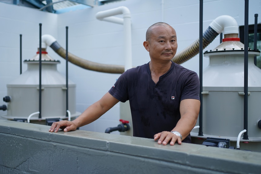 Chinese man, casually dressed, standing in an industrial space with large water tanks and piping in the background. 