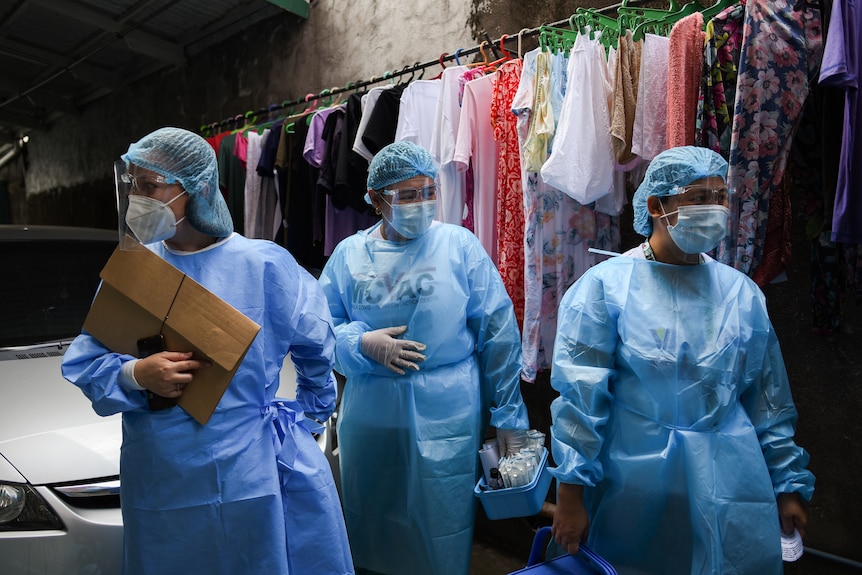 Three women in full PPE stand next to a row of hanging clothes 
