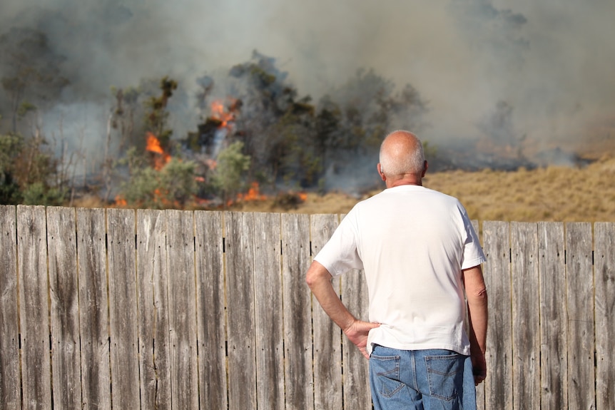 A man in a white shirt looks over a fence with flames and smoke in the background.