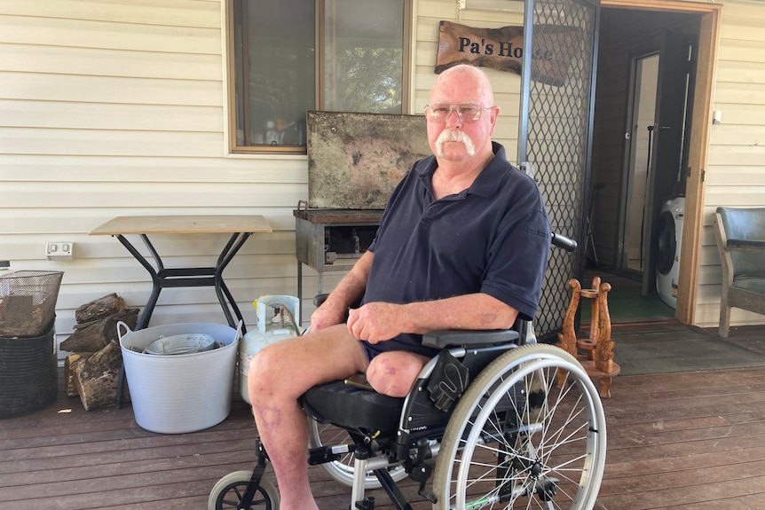 A bald man, grey handlebar moustache, blue t-shirt, glasses, sits in a wheel chair in a weatherboard house, Pa's house sign.
