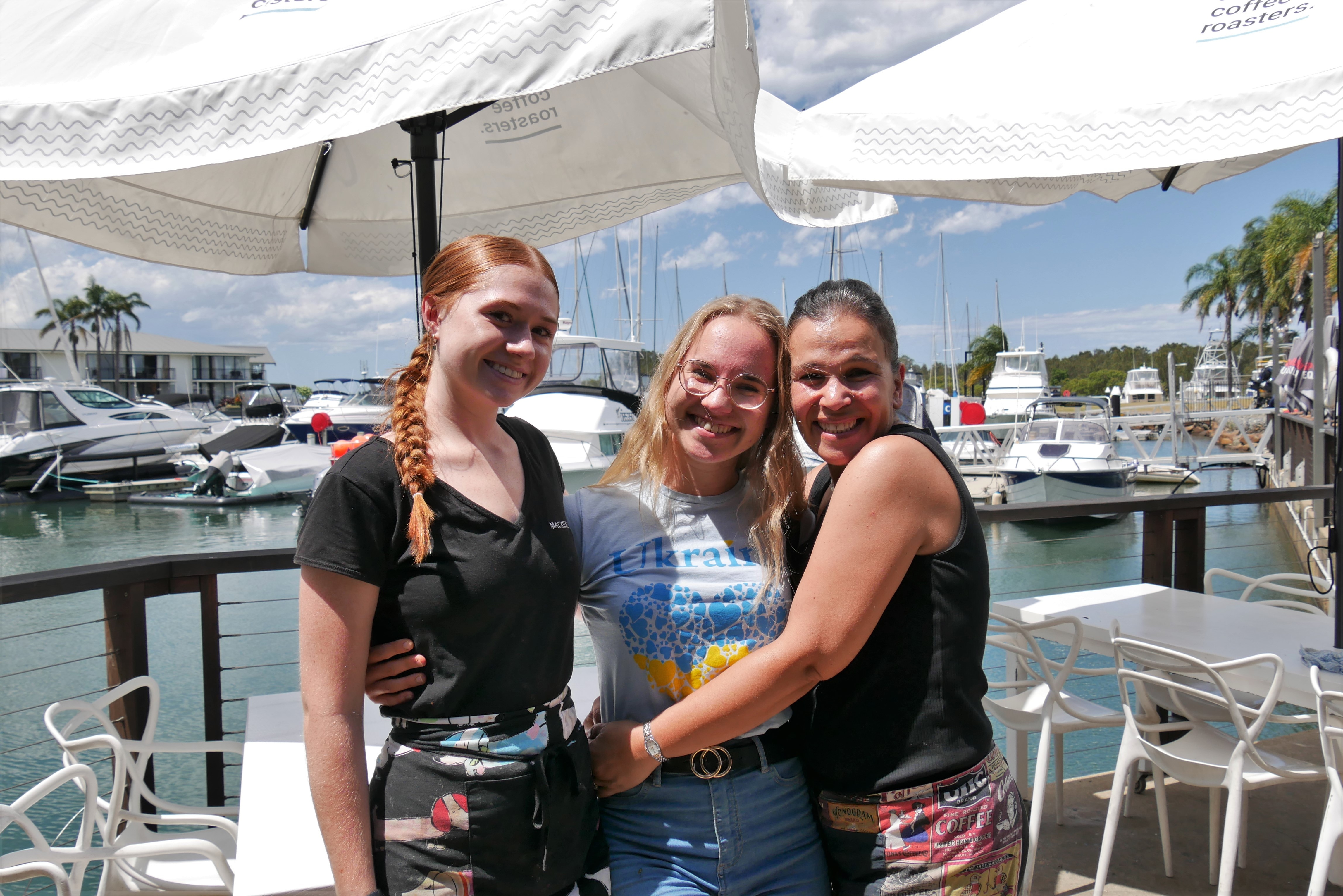 Three women smile at the camera with their arms around one another. A boat harbour is in the background. 