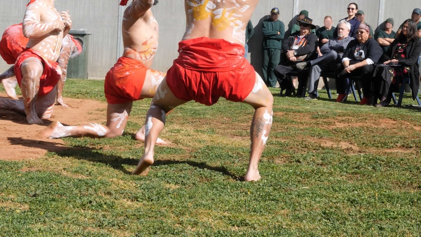 Four men with white marking painted on their bodies, wearing red loincloths, dance before a small crowd.