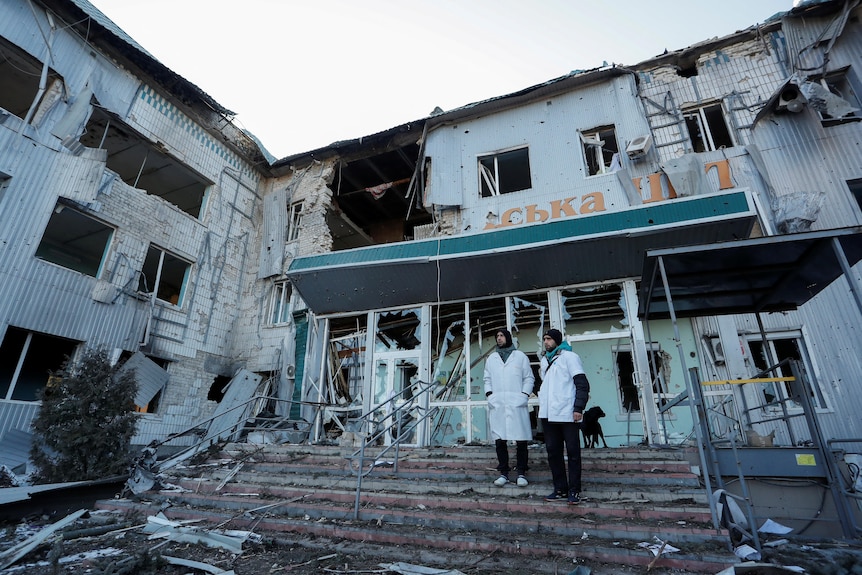 Two medical workers stand on the front steps of a hospital building, which has been badly damaged, walls and windows missing.