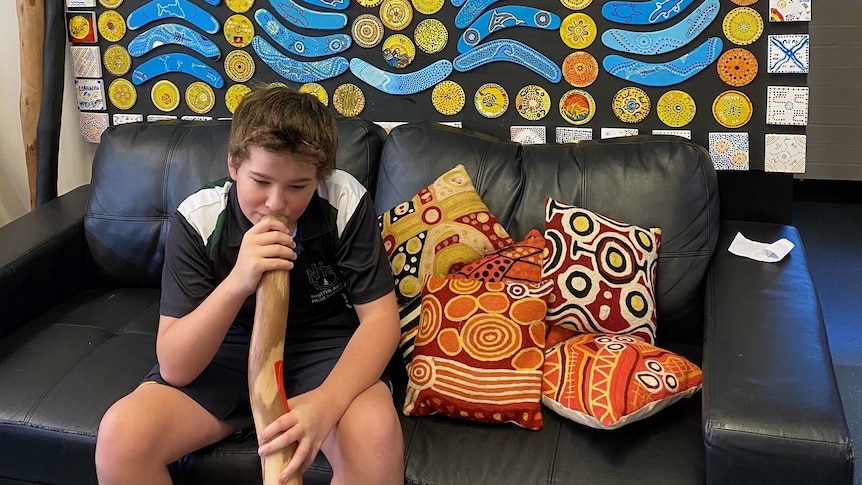 Archie playing a didgeridoo, sitting on a couch strewn with cushions printed with Indigenous art, in front of a board of art.