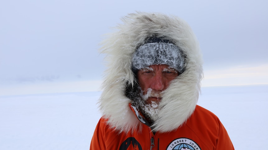 A man in a snow jacket stares into the camera with snow stuck to his beard and face