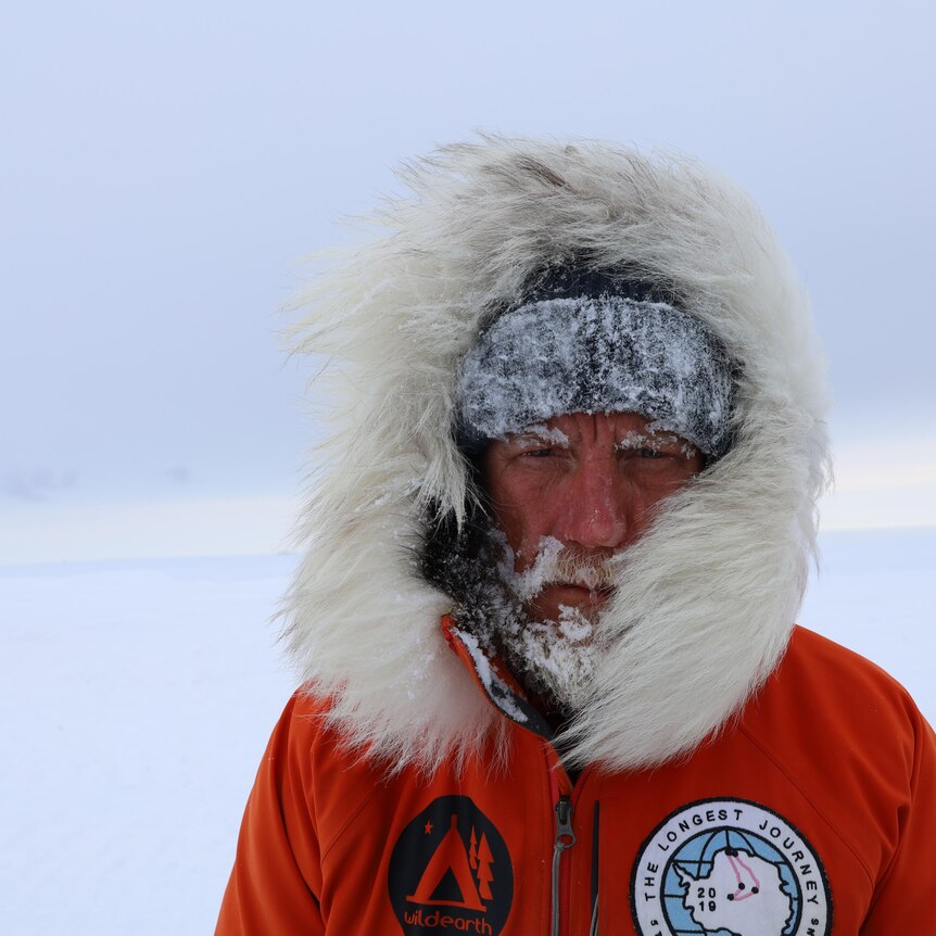 A man in a snow jacket stares into the camera with snow stuck to his beard and face
