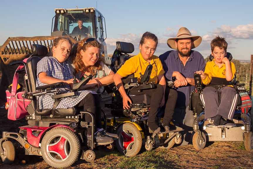 A family including three children in power wheelchairs with a mother and father on a dirt patch with a digger behind them