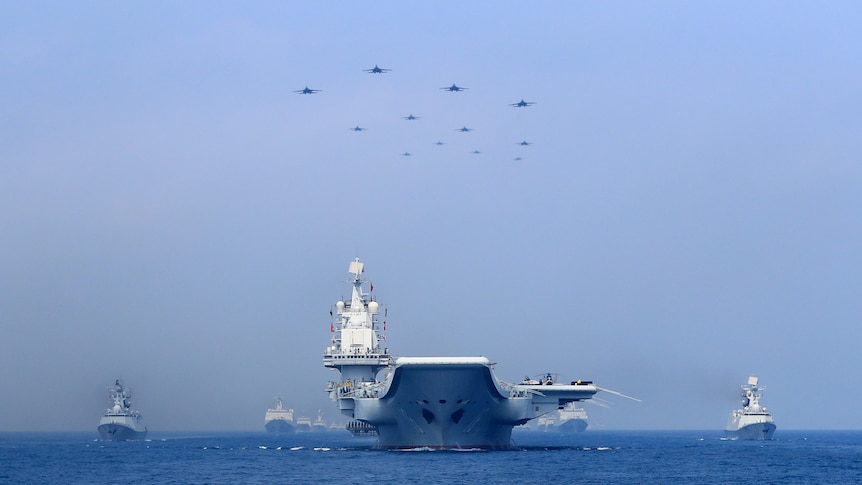 A very big warship, surrounded by smaller ships, with planes flying in formation overhead