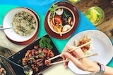 Bowls of tom yum soup, fried rice and marinated meat, with a hand using chopsticks to eat the Thai food.