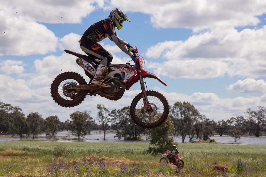 A man airborne on a motorbike in a paddock with a flooded river in the background