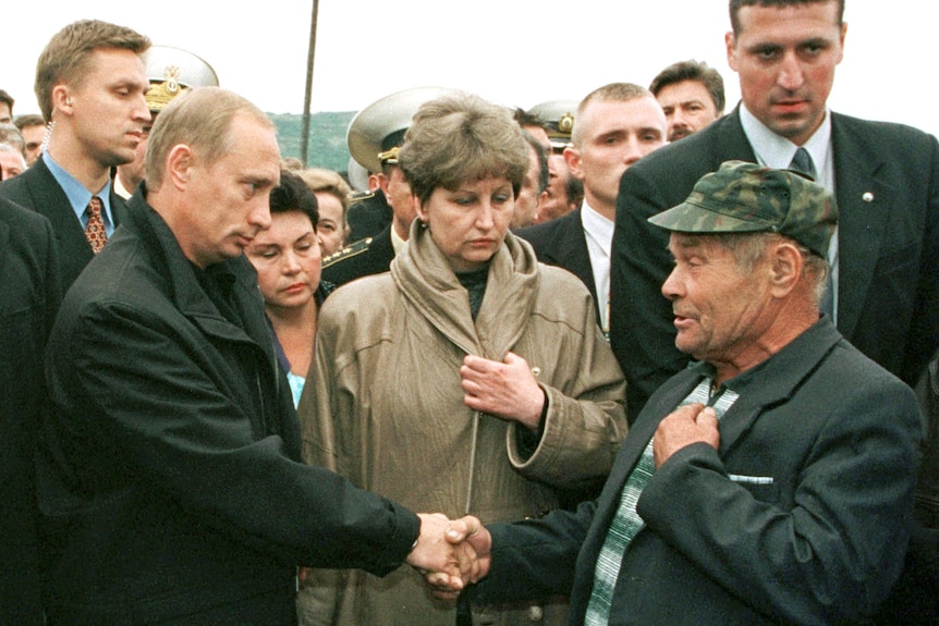 Vladimir Putin shaking hands with a man while a crowd watches 