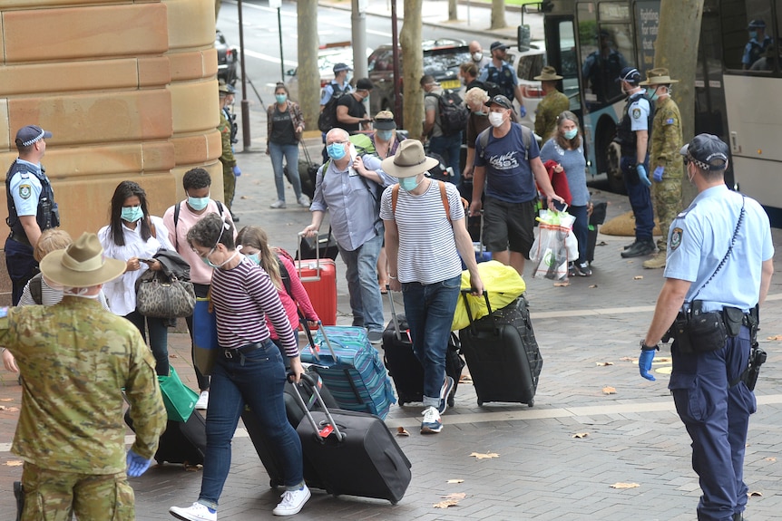 people wearing masks and pulling bags with wheels walk on the footpath