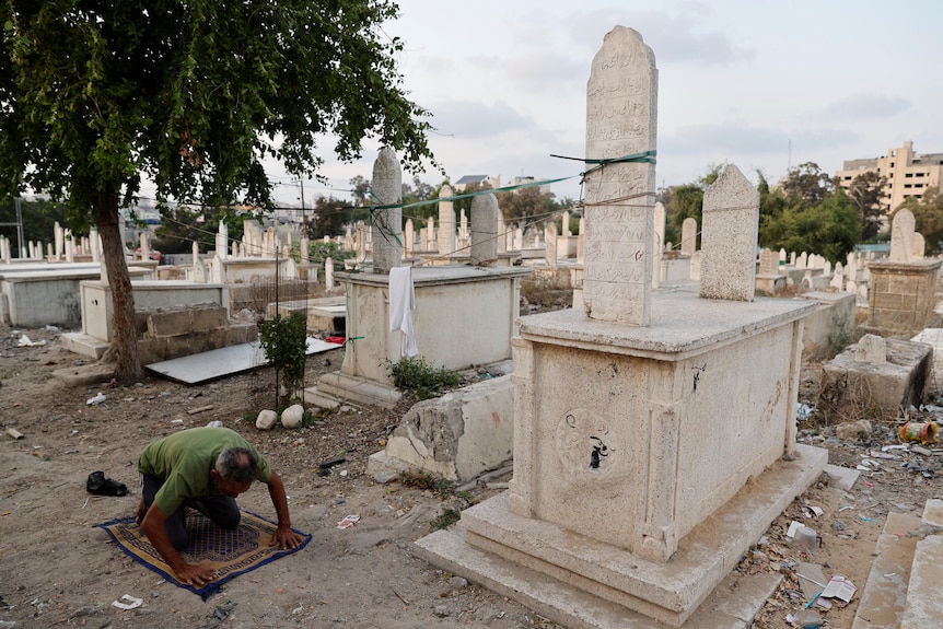 A man kneels and prays on a mat in the middle of a crumbling cemetery. 