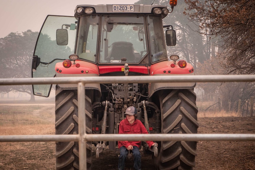 Will Kippel wears a a red shirt and cap, he sits in the middle of two big wheels of a red tractor.