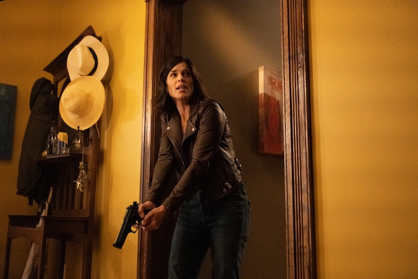 A woman in her 40s wearing a leather jacket stands at the ready in the doorway of a house, looking distressed and  holding a gun