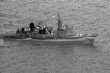 A black and white image showing an aerial view of a military ship on a calm ocean.