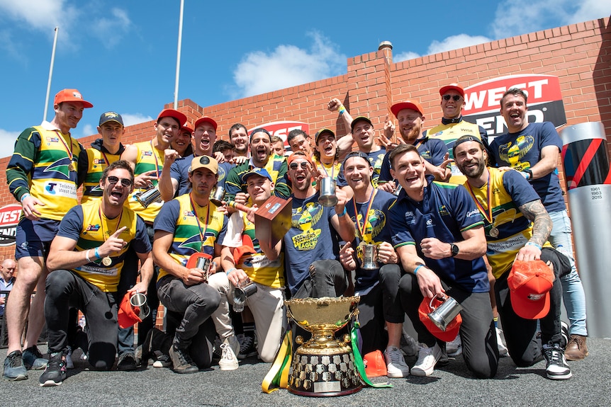 A football team huddle around a premiership cup and celebrate with a brick building behind them.