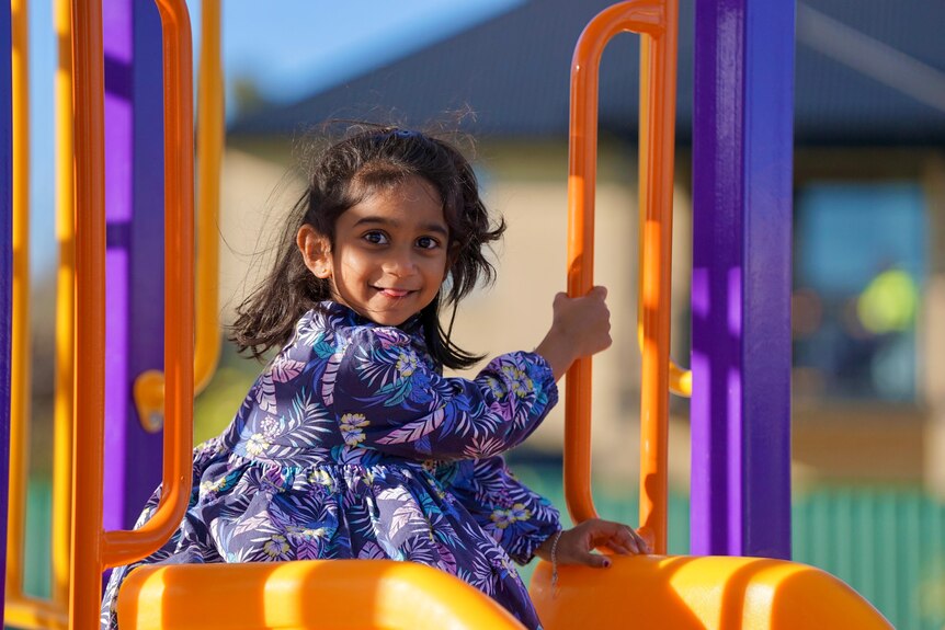 A little girl wearing purple floral smiles big while sitting atop a playground slide