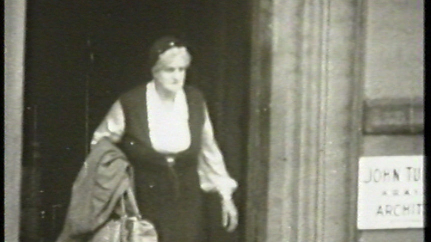 Black and white image of middle aged woman walking out of a building entrance.