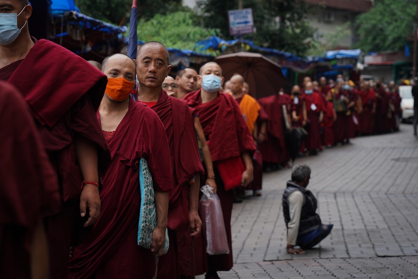 A row of monks in their trademark red and orange robes line up, a person with no legs sits nearby