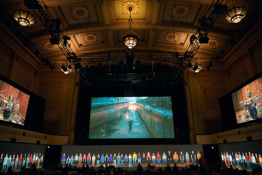 A row of people line a stage in Melbourne's Town Hall, with images projected on a screen behind them.