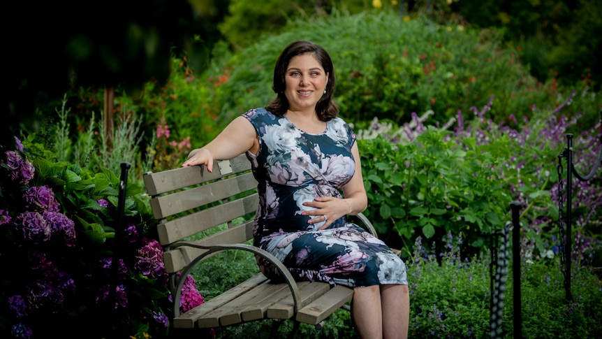 Nas sits on a park bench surrounded by lush green plants and purple flowers, holding her baby bump with one hand and smiling.
